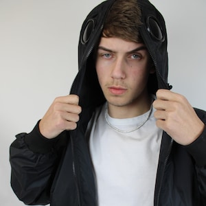 May include: A black hooded jacket with a zipper closure and two circular eyelet details on the hood. The jacket is made of a lightweight fabric and has a relaxed fit.