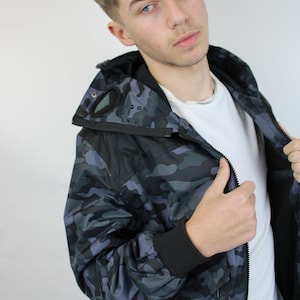 May include: A black and gray camouflage jacket with a zipper closure and a hood. The jacket has a black eye patch on the hood and a black strap on the left side.
