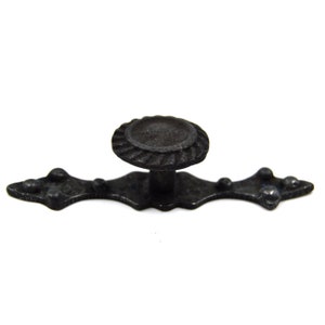 May include: Black metal cabinet knob with a decorative design. The knob has a round, raised center and a flat, rectangular base.