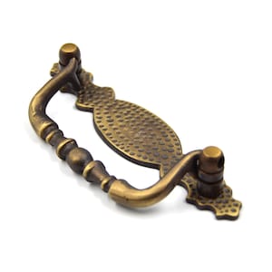 Puede incluir: Tirador de armario de latón antiguo con una placa ovalada texturizada y un asa curva. El tirador tiene un acabado desgastado y un diseño clásico.