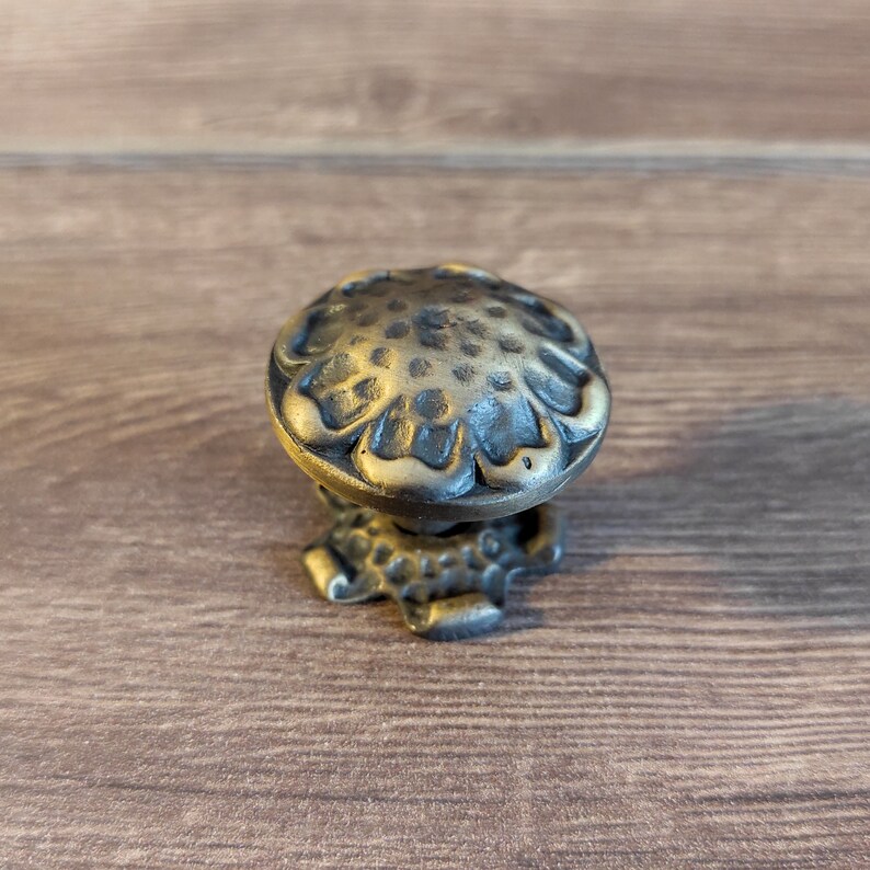 Puede incluir: Un pomo de armario de metal dorado y negro con un dise&ntilde;o floral. El pomo tiene una superficie texturizada y una forma redondeada.