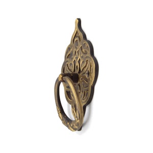 May include: An ornate metal ring pull handle in a gold tone, featuring intricate floral designs. The handle is attached to a decorative plate with a similar design.