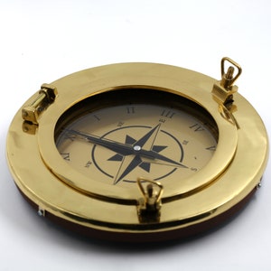 Könnte beinhalten: Ein Kompass aus Messing mit römischen Ziffern und einer Kompassrose. Der Kompass ist in einen runden Messingrahmen mit einer Holzrückseite eingefasst.