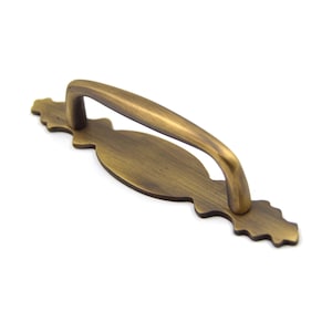 May include: An antique brass cabinet pull with an ornate design. The pull has a curved handle and a decorative plate with a scalloped edge.