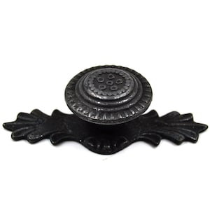 Puede incluir: Pomo de armario de metal negro con un diseño decorativo y ornamentado. El pomo tiene un centro redondo y elevado con un patrón circular y una base plana más pequeña.