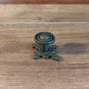 May include: A dark grey metal cabinet knob with a decorative, ornate design. The knob has a round top and a base with a starburst pattern.