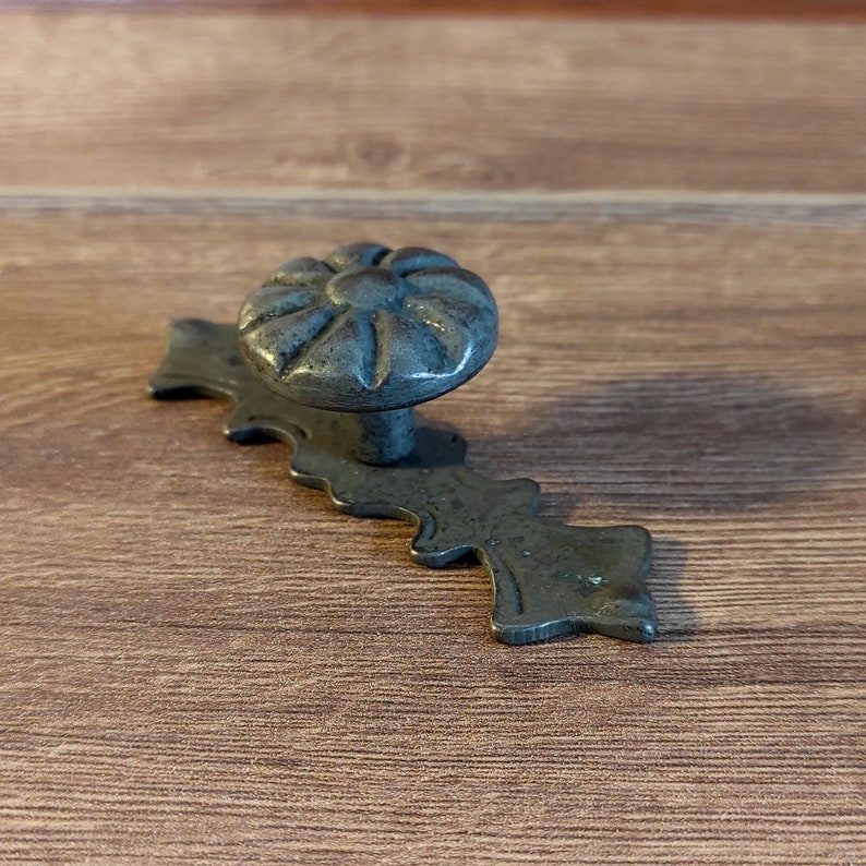 Pu&ograve; includere: Una maniglia per cassetto in metallo nero con un bordo decorativo a festoni e un pomo a forma di fiore.