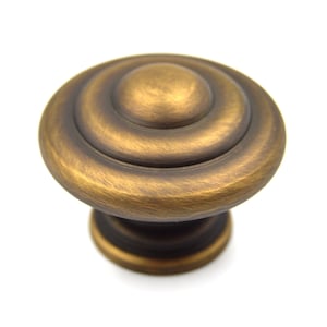 May include: A round, antique brass cabinet knob with a raised, circular design. The knob has a slightly textured finish.