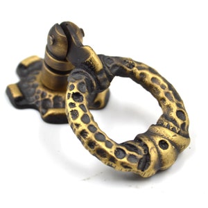 Op de afbeelding: Een messing ringgreep voor een kastdeur met een gestructureerd oppervlak. De greep heeft een ronde ring en een basis met een decoratief ontwerp.