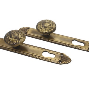 Op de afbeelding: Twee antieke messing deurgrepen met sierlijke bloemmotieven. Elke greep heeft een ronde knop met een sleutelgatplaat.