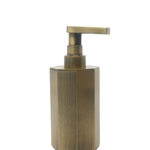 May include: A gold-toned soap dispenser with a hexagonal base and a pump top. The dispenser has a brushed metal finish.