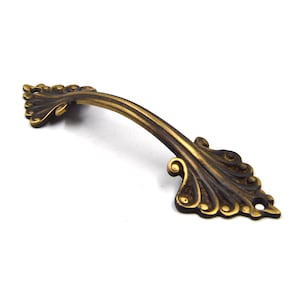 Puede incluir: Un tirador de armario de latón decorativo con un diseño floral ornamentado. El tirador tiene una forma curva y un acabado oscuro y antiguo.