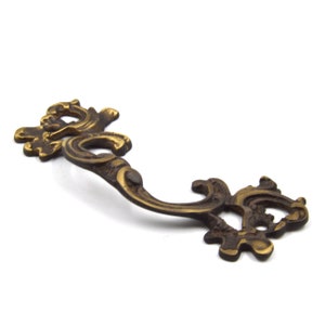 May include: A gold-toned metal cabinet pull with an ornate, scroll-like design.