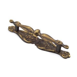 May include: Ornate brass drawer pull with a decorative floral design. The pull has a dark brown patina and is approximately 4 inches long.