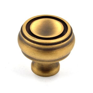 Puede incluir: Un pomo de armario redondo de color dorado con un diseño circular elevado. El pomo tiene un acabado liso y cepillado.