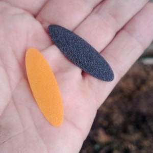 Puede incluir: Dos púas de guitarra, una naranja y otra negra, se muestran en una mano. Las púas son de forma ovalada y tienen una superficie texturizada.