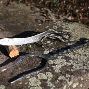 Könnte beinhalten: Zwei handgeschmiedete Metallwerkzeuge, ein Messer und eine Gabel, mit gedrehten Griffen, die auf einer grauen Steinoberfläche mit Flechten liegen.