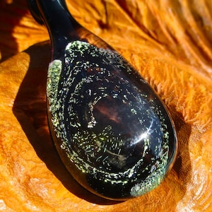 May include: A dark glass teardrop pendant with green and gold glitter embedded inside. The pendant is resting on a wooden surface.