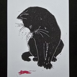 Könnte beinhalten: Ein schwarz-weißer Linoldruck auf Papier, der eine schwarze Katze zeigt, die sitzt und auf eine kleine, leuchtend rosa Maus hinunterblickt. Die Katze hat sichtbare weiße Schnurrhaare und strukturierte Fellzeichnungen. Die Signatur steht unten.