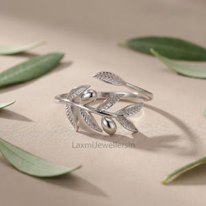 May include: A silver-colored ring in the shape of an olive branch, with leaves and olives. The ring is surrounded by green olive leaves on a neutral background. The text "LaxmiJewellersIn" is visible.
