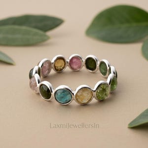 Op de afbeelding: Een zilveren ring met een reeks kleine, ronde, veelkleurige edelstenen in roze, gele, groene en blauwe tinten. De stenen zijn gezet in individuele zilveren zettingen die in een doorlopende cirkel zijn gerangschikt. De tekst LaxmijewellersIn is zichtbaar.