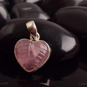 May include: A heart-shaped pendant with a pink gemstone, set in a gold-colored metal. The gemstone has a carved leaf-like design. The pendant is displayed on a bed of black, polished stones.