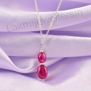 Puede incluir: Un collar de plata con cadena y dos piedras preciosas ovaladas de color rosa intenso. Las piedras están apiladas, con una pequeña cuenta de plata entre ellas. El collar se exhibe sobre una tela lavanda.