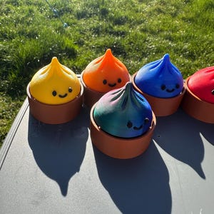 Puede incluir: Una colección de objetos coloridos con forma de dumpling sonriente en amarillo, naranja, azul, rojo y una variante multicolor. Cada dumpling está en un recipiente marrón y circular. Los objetos están dispuestos sobre una superficie gris, con un fondo de hierba.