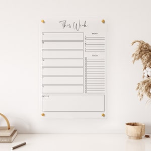 May include: A clear acrylic weekly planner with gold push pins. The planner has sections for each day of the week, a menu section, a to-do list, and a notes section.