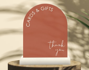 May include: A clear acrylic sign with a rounded top and a terracotta background. The sign says "Cards & Gifts" at the top and "Thank You" at the bottom in white lettering.