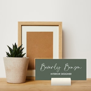May include: A green acrylic sign with white text that reads "Beverly Benson INTERIOR DESIGNER". The sign is on a white stand and is sitting on a wooden surface. There is a potted succulent plant and a wooden picture frame in the background.