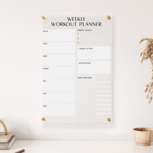 May include: A white acrylic weekly workout planner with gold push pins. The planner has sections for weekly goals, how you want to feel, motivation, and a habit tracker.