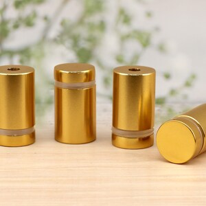 May include: Four gold-colored metal standoffs with a clear plastic insert. The standoffs are arranged in a row on a light wood surface.