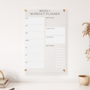 May include: A white acrylic weekly workout planner with gold push pins. The planner has sections for weekly goals, how you want to feel, motivation, and a habit tracker.