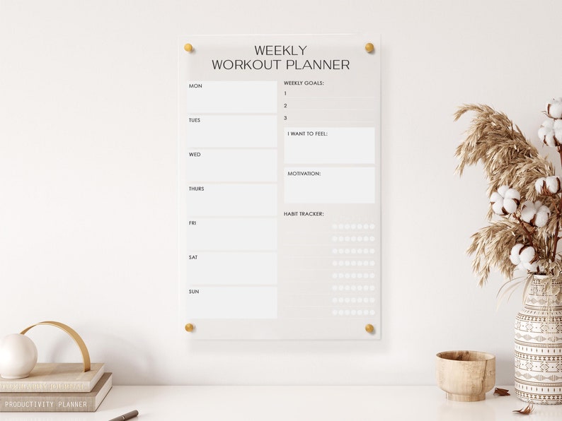 May include: A white acrylic weekly workout planner with gold push pins. The planner has sections for weekly goals, how you want to feel, motivation, and a habit tracker.