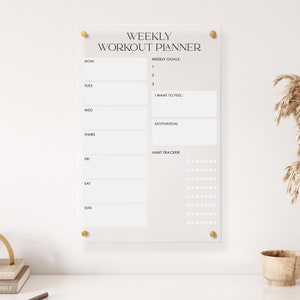 May include: A white acrylic weekly workout planner with gold accents. The planner includes sections for weekly goals, motivation, and a habit tracker. The planner is hanging on a white wall.
