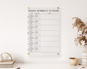 May include: A weekly workout planner on a white background with a black and white design. The planner has columns for the days of the week, focus, intensity, and outline. The planner is hung on a wall with gold push pins.