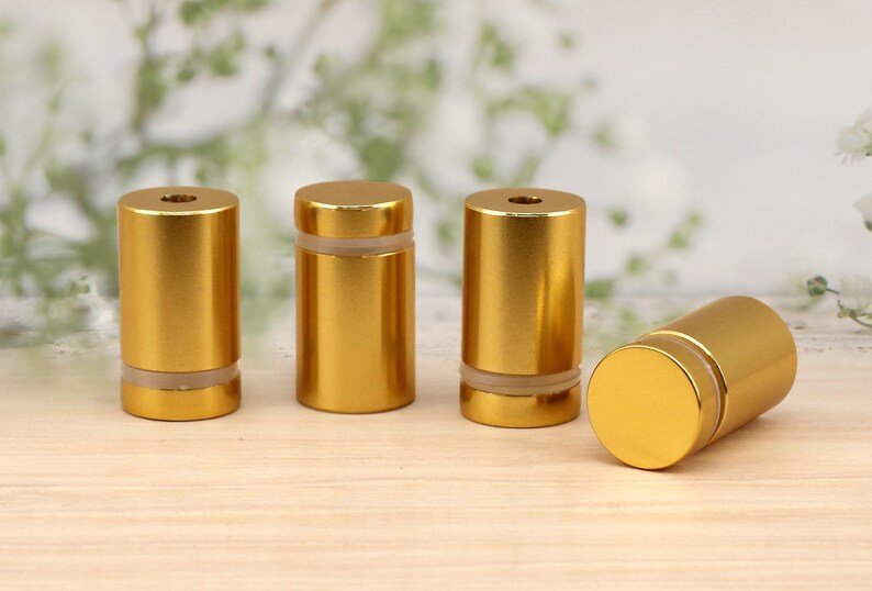 May include: Four gold-colored metal standoffs with a clear plastic insert. The standoffs are arranged in a row on a light wood surface.