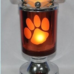 Könnte beinhalten: Ein dekorativer elektrischer Ölverdampfer mit Pfotenabdruck-Design. Der Verdampfer hat einen roten Glaskörper mit einem Chromsockel und -deckel. Der Deckel hat eine wellenförmige, orangefarbene Glasschale. Ca. 20 cm hoch.