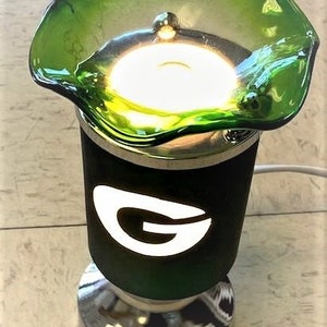 May include: A green glass oil diffuser with a white "G" logo on the front. The diffuser has a chrome base and a green glass top with a light bulb inside.