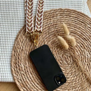 Könnte beinhalten: Ein schwarzes Handy-Case mit einer goldenen Kette und einem braunen und weißen gemusterten Handy-Band. Das Handy-Case und das Band liegen auf einer geflochtenen Strohmatte.