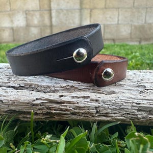 May include: Two leather bracelets, one black and one brown, are displayed. Each bracelet has a silver-colored snap closure. The bracelets are resting on a weathered wooden surface, with green grass and a brick wall in the background.