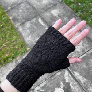 Puede incluir: Un guante negro sin dedos hecho de punto. El guante cubre la muñeca y se extiende hasta los nudillos, dejando los dedos expuestos. La textura de punto es visible, y el guante parece estar hecho de un hilo suave y cálido.