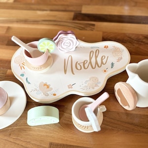 May include: A wooden tea set with a cream-colored base and pastel accents. The set includes a tray with the name "Noelle", a teapot, a cupcake, and other tea party accessories. The set is decorated with floral designs.