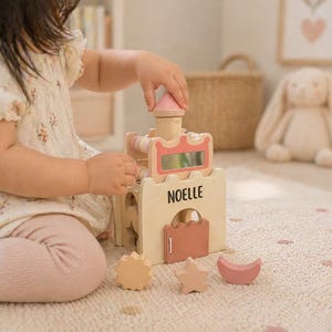 May include: Wooden toy castle with pink and natural wood accents. The castle features a pink roof, a decorative pink frame, and the name "NOELLE" printed on the front. Includes a sun, star, and moon-shaped blocks.