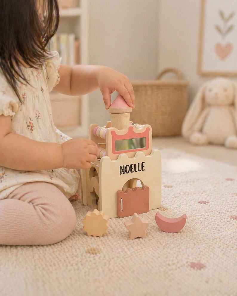 May include: Wooden toy castle with pink and natural wood accents. The castle features a pink roof, a decorative pink frame, and the name "NOELLE" printed on the front. Includes a sun, star, and moon-shaped blocks.