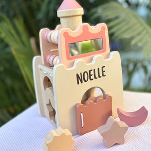 May include: Wooden toy castle with the name "NOELLE" printed on the front. The castle features a pink and beige color scheme, with a tower, a mirror, and various shape sorter pieces, including a star, sun, and crescent moon.