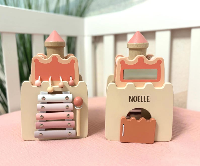 May include: Two wooden toy castles with peach and light brown accents. One castle features a xylophone with pink and white keys, the other has the name "NOELLE" printed on it. Both have a pink turret.