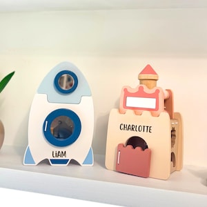 May include: Two wooden children's toys: a rocket ship and a castle. The rocket is white with blue and light blue accents, and the name "LIAM" is printed on it. The castle is light brown with pink accents, and the name "CHARLOTTE" is printed on it.