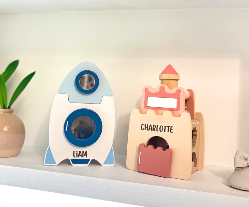 May include: Two wooden children's toys: a rocket ship and a castle. The rocket is white with blue and light blue accents, and the name "LIAM" is printed on it. The castle is light brown with pink accents, and the name "CHARLOTTE" is printed on it.